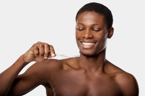 Shirtless African American man in a studio, brushing his teeth with a toothbrush against a white background. — Stock Photo
