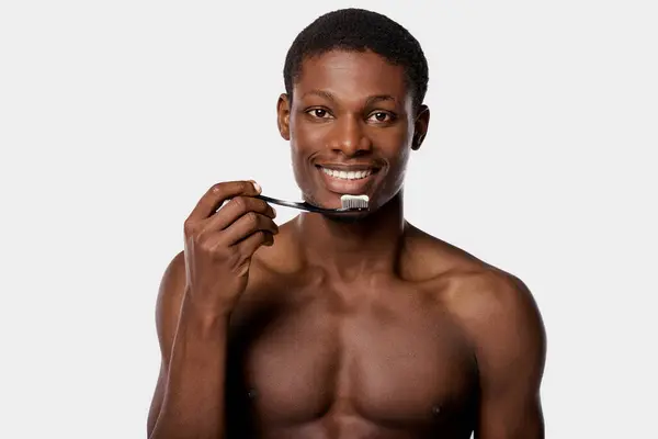 A shirtless African American man brushing his teeth with a toothbrush in a white studio background. — Stock Photo