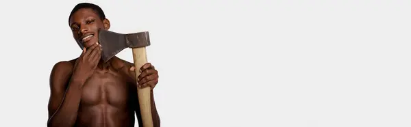 Un afroamericano senza maglietta tiene una grande ascia nella mano destra sullo sfondo di uno studio bianco. — Foto stock
