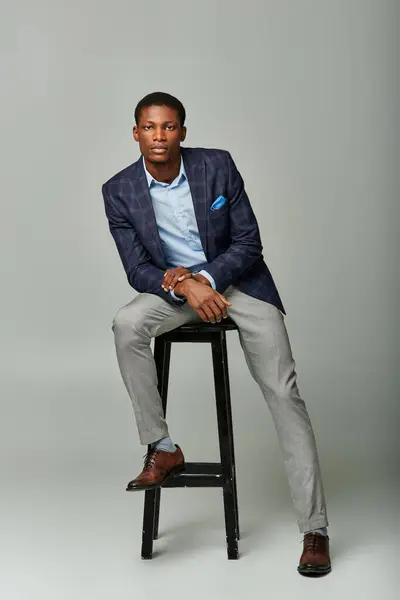Bello giovane uomo d'affari in giacca a scacchi, seduto con fiducia sullo sgabello sullo sfondo grigio. — Foto stock