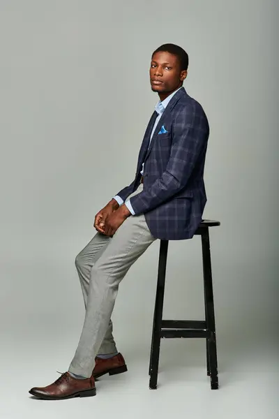 Ein gutaussehender afroamerikanischer Mann in kariertem Blazer und formeller Kleidung sitzt selbstbewusst auf einem Hocker vor grauem Hintergrund.. — Stockfoto