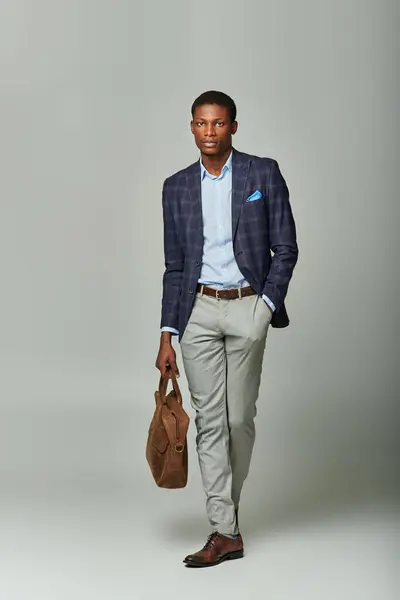 An African American man in a stylish suit and tie, confidently holding a bag on a grey background. — Stock Photo