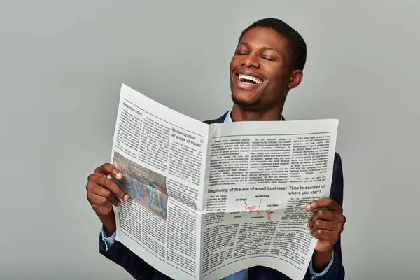 A handsome African American man in a checkered blazer reading a newspaper and laughing heartily. — Stock Photo