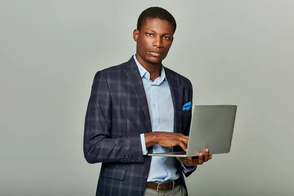 African American businessman in checkered blazer confidently holds a laptop on grey background. — Stock Photo