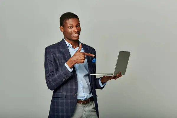 A stylish African American businessman in a checkered suit holding a laptop against a sleek grey background. — Stock Photo