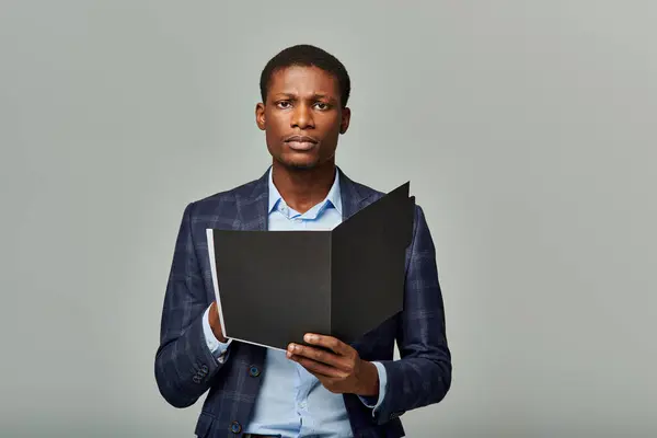 Homme d'affaires afro-américain élégant dans un blazer à carreaux tient avec confiance un dossier dans un cadre gris élégant. — Photo de stock