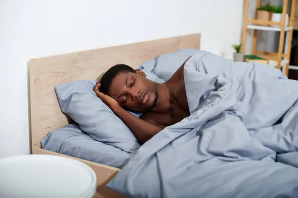 A young African American man peacefully sleeping in bed under a blue blanket, beginning to wake up in the morning. — Stock Photo