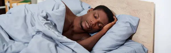 Ein junger afroamerikanischer Mann schläft friedlich im Bett unter einer kuscheligen blauen Decke, bereit, am Morgen aufzuwachen. — Stockfoto