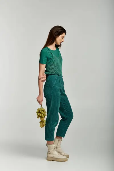 A young woman in a green outfit holds a bunch of grapes in a white studio. — Stock Photo