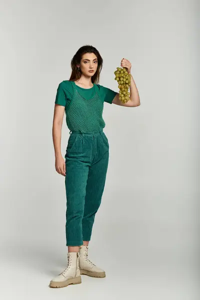 A young woman wearing green clothing holds a bunch of grapes in a studio setting. — Stock Photo