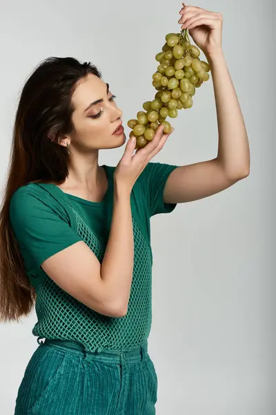 A young woman in a green shirt and pants smells a bunch of grapes. — Stock Photo