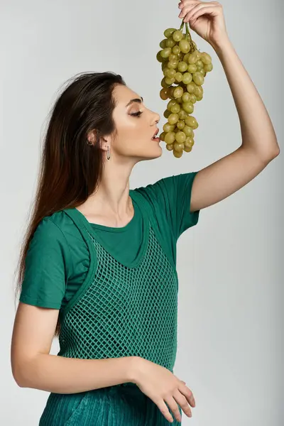 A young woman in a green outfit holds a bunch of grapes to her lips. — Stock Photo