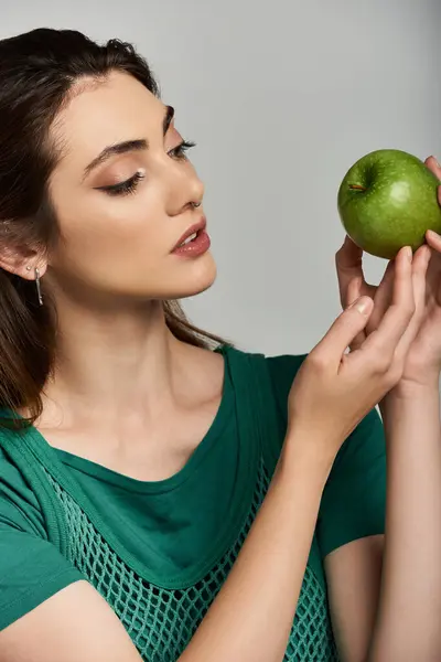 A beautiful woman, dressed in green, gazes at a green apple held in her hand. — Stock Photo