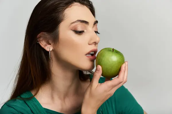 A young woman in a green shirt holds a green apple to her lips, about to take a bite. — Stock Photo
