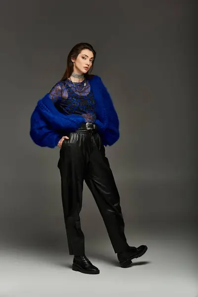 Young woman in blue cardigan, leather pants, and black boots poses against grey background. — Stock Photo