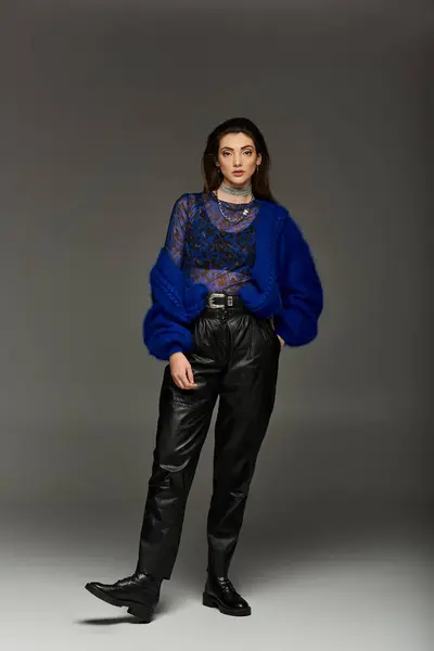 A young woman in a blue cardigan and black leather pants stands confidently in a studio setting. — Stock Photo