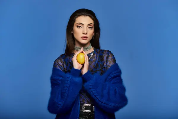 A young woman in a blue sweater holds a lemon in front of a blue background. — Stock Photo