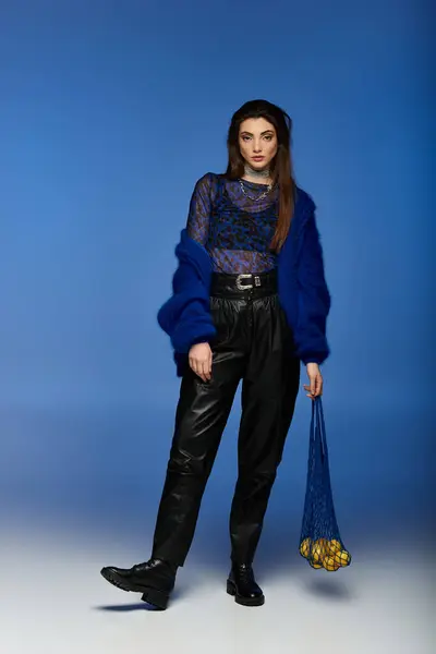 A woman in a blue fuzzy cardigan and black pants stands in front of a bright blue background. — Stock Photo