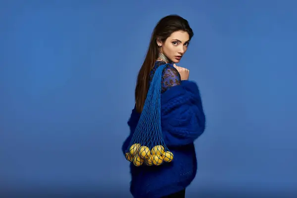 Young woman in blue cardigan holding yellow fruit in mesh bag on blue background. — Stock Photo