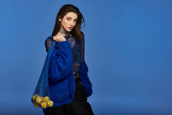 Woman in blue sweater holds mesh bag with lemons on blue backdrop. — Stock Photo