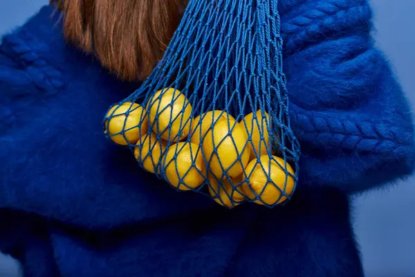A woman in a blue fuzzy sweater carries a net bag filled with lemons. — Stock Photo