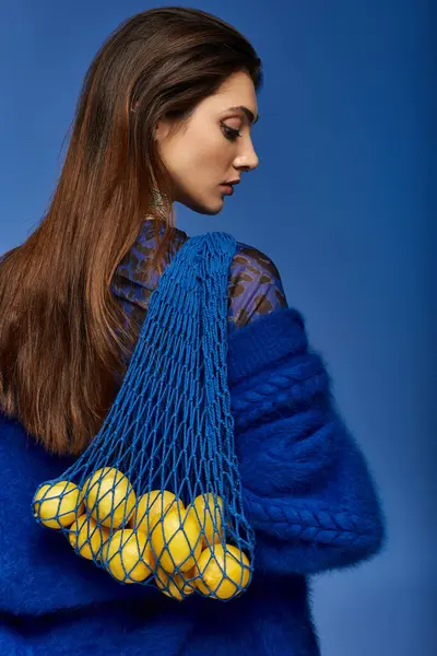 A young woman in a blue sweater carries a net bag filled with lemons. — Stock Photo