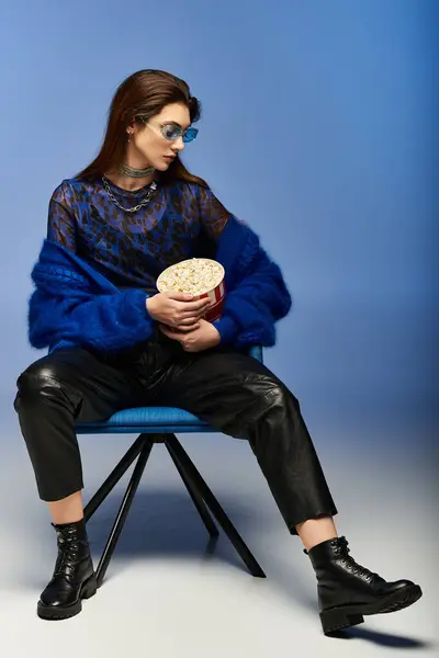 Eine junge Frau in schicker Kleidung sitzt auf einem blauen Stuhl und hält einen Behälter mit Popcorn in der Hand. — Stockfoto
