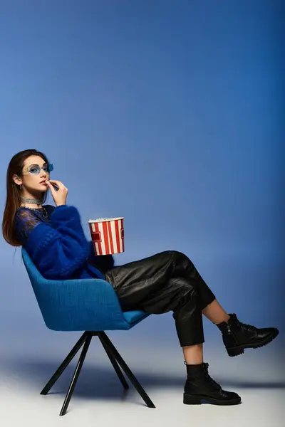 Woman in elegant attire sits in blue chair with popcorn, looking away against a blue background. — Stock Photo