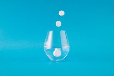 Parlak mavi bir arka planda, bir bardak suda çözünen coşkulu bir tablet. Temiz sıvı ve fokurdayan tablet canlandırıcı ve temiz bir görsel yaratıyor.,