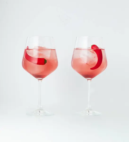 stock image two wine glasses filled with rose-colored cocktails, each garnished with a chili pepper and ice cubes, set against a white background. The combination of spicy chili
