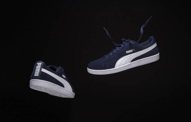 Puma spor ayakkabılarının siyah arka planda asılı olduğu dinamik bir stüdyo çekimi beyaz çizgili lacivert tasarımı vurguluyor. Modern spor ayakkabılarını, stilini ve markasını sergilemek için mükemmel..