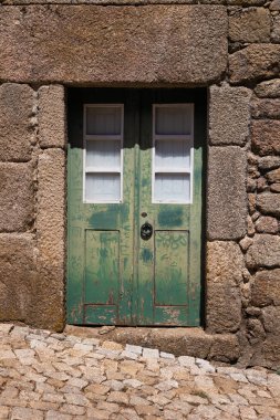 Portekiz, Monsanto 'da kırsal taştan bir duvarda aşınmış yeşil bir kapı. Eski ahşap ve yıpranmış boyalar tarihi çağrıştırırken, kaldırım taşı caddesi bu ortaçağ köyünün eski dünya cazibesini artırıyor..