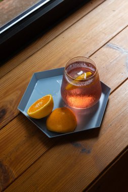 A refreshing citrus cocktail served with fresh orange halves, placed on a geometric tray atop a wooden table. The natural lighting and condensation on the glass add a cozy, relaxed feel. clipart