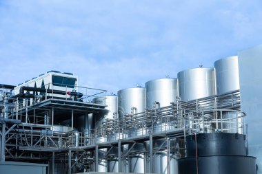 Parlak mavi gökyüzü altında paslanmaz çelik depolama tankları, karmaşık boruları ve modern makineleri olan endüstriyel bir fabrikanın dış görünüşü. Endüstriyel mühendislik ve imalatın mükemmel bir temsili.