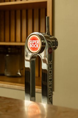 Modern bir bar ortamında Super Bock bira fıçısına yakın çekim. Cilalı krom kaplama ve marka logosu öne çıkıyor, kaliteli bira servisi ve işçiliğin özünü yakalıyor..