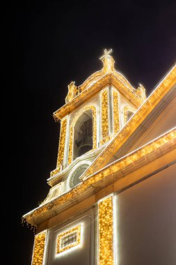 Night view of Igreja do Livramento in Mafra, Portugal, illuminated with festive golden lights. The glowing church facade and bell tower stand out beautifully against the dark night sky. clipart