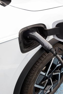 Şarj olan elektrikli bir araca yakın çekim. Beyaz araba, çevre dostu ulaşım eğilimlerini vurgulayan siyah bir kabloyla şarj istasyonuna bağlı..