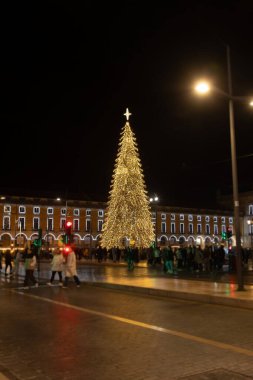 A beautifully illuminated Christmas tree stands tall in a city square at night, surrounded by crowds enjoying the festive atmosphere. The bright lights and holiday decorations create a magical urban in Lisbon clipart