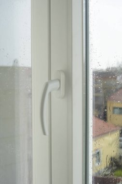 Camdan damlayan yağmur damlalarıyla dolu modern beyaz bir pencere kolunun yakın çekimi bulanık bir şehir arka planını çerçeveliyor. Sahne, kapalı alanda yağmurlu bir günün sükunetini ve dinginliğini yakalar..