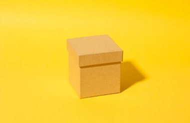 Kapaklı basit bir karton kutu parlak sarı bir arkaplanda duruyor. Minimalist kompozisyon, temiz çizgiler ve yumuşak gölgeler modern bir estetik yaratıyor, merak ve gizem uyandırıyor..