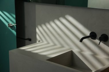 Minimalist beton lavabosu ve yumuşak doğal ışıkla aydınlatılan parlak siyah duvarlı musluğu olan modern bir banyo. Perdelerin gölgeleri çarpıcı bir desen oluşturur, çağdaş uzaya derinlik ve zarafet ekler..