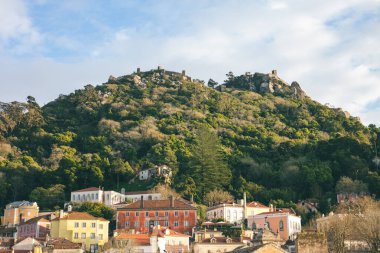 Portekiz 'in Sintra kentinin yeşil tepelerinin üzerindeki Fas Kalesi' nin göz kamaştırıcı manzarası. Önünde renkli tarihi evler var. Bu pitoresk manzara ortaçağ tarihi, doğası ve büyüleyici mimarisini harmanlıyor..