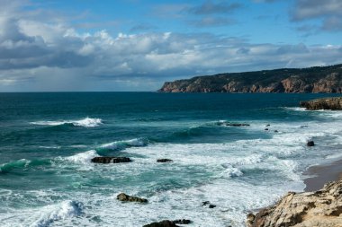 A breathtaking view of Guincho Beach in Cascais, Portugal, where powerful Atlantic waves crash against the rugged coastline. The turquoise sea, rocky cliffs, and dramatic sky create a stunning coastal scene.