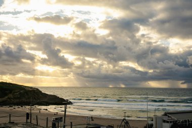 Praia das Mas, Sintra 'da nefes kesen bir gün batımı. Atlantik Okyanusu' nda altın güneş ışınlarını filtreleyen dramatik bulutlar. Dalgalar kıyıya çarpıyor, sakin ama güçlü bir kıyı manzarası yaratıyor..