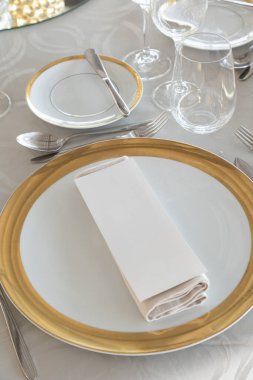 Beyaz ve altın tabaklı, gümüş çatal bıçaklı ve kristal gözlüklü resmi bir etkinlik için şık bir masa. Katlanmış bir peçete ve boş bir menü kartı sofistike yemek düzenine ince bir dokunuş ekler