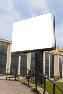 Büyük boş bir ilan panosu modern cam binalara karşı kentsel bir ortamda duruyor. Reklamlar için mükemmel bir yer sunuyor. Boş beyaz alan onu pazarlama modelleri ve promosyon tasarımları için ideal hale getiriyor.