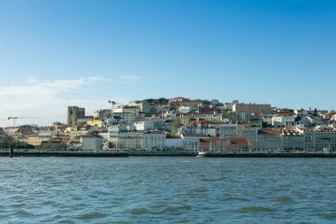 Lizbon 'un tarihi Alfama bölgesinin, Tagus Nehri' ndeki bir tekneden, berrak mavi gökyüzü altında geleneksel mimari ve renkli binalar sergilenen pitoresk bir manzarası..