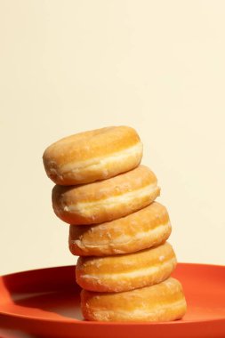 Beş altın kaplamalı donut yığını canlı turuncu bir tabağa yaslanır ve yumuşak gölgeler bırakır. Minimalist bej arka plan bu tatlı düzeneğin sıcak tonlarını ve yumuşak dokularını vurgular.