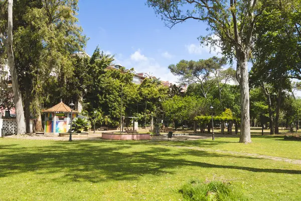 Portekiz 'in Oeiras kentindeki bir halk parkında güneşli bir gün, büyük yeşil bir çimenlik, uzun ağaçlar ve renkli bir büfeyle. Huzurlu kentsel manzara rahatlama, dinlenme veya turizm görselleri için ideal.