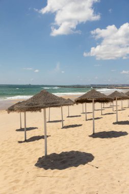 Portekiz 'in Costa da Caparica şehrinin altın kumlarına gölge düşüren saman plajı şemsiyeleri. Parlak bir gökyüzü altında sakin bir deniz manzarası, rahatlama ve yaz tatili görüntüsü için mükemmel..